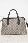 A Gucci handbag in beige and brown GG Supreme canvas with black leather handles and trim, displayed on a light marble surface against a white background.