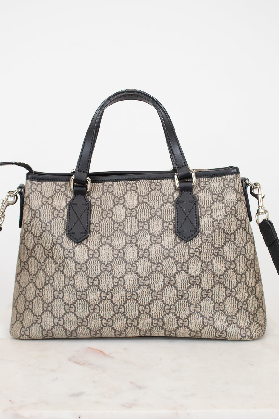 A Gucci handbag in beige and brown GG Supreme canvas with black leather handles and trim, displayed on a light marble surface against a white background.