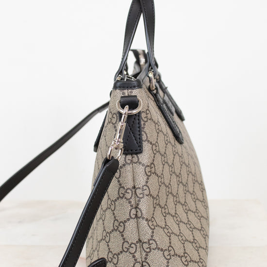 A beige and brown GG Supreme canvas handbag with black leather trim, handles, and shoulder strap, shown from a side profile view on a light background.