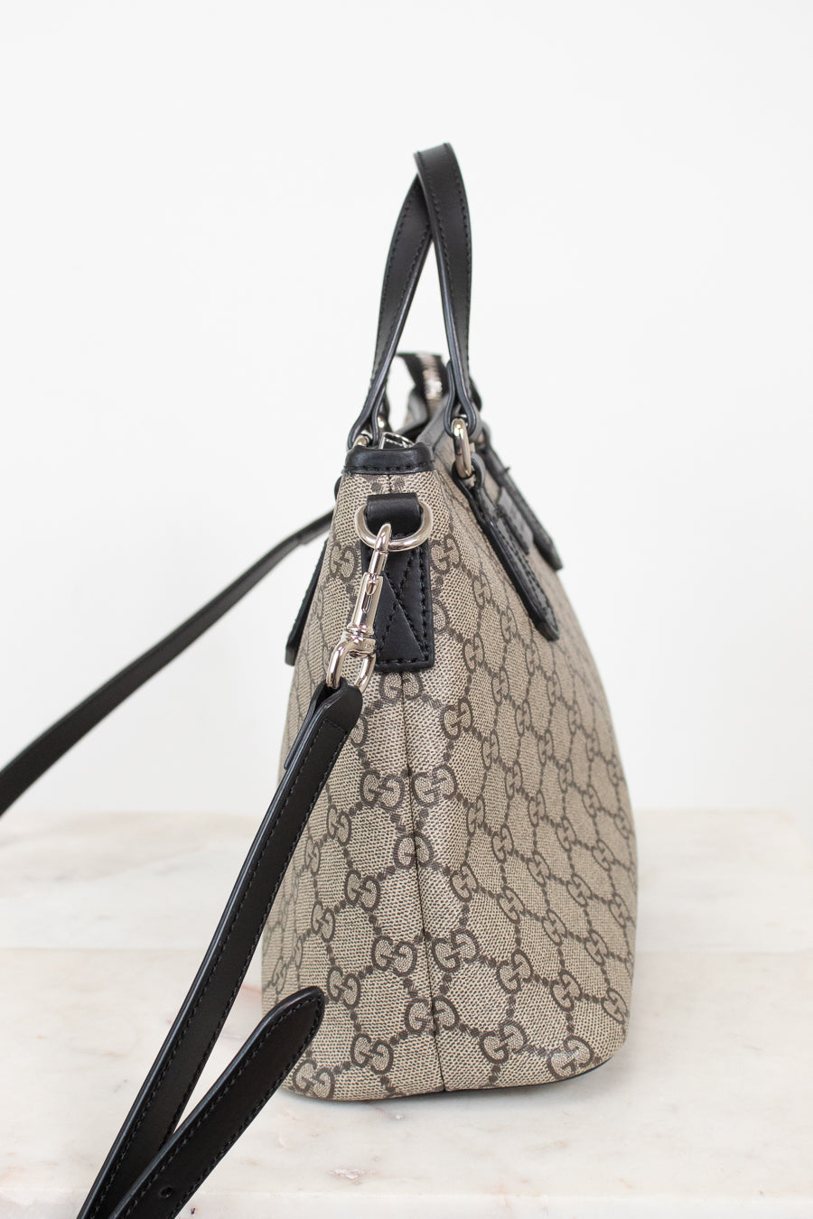 A beige and brown GG Supreme canvas handbag with black leather trim, handles, and shoulder strap, shown from a side profile view on a light background.