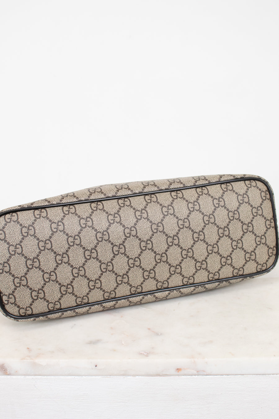 A Gucci monogram canvas bottom view or small bag with a beige and brown GG pattern, resting on a white marble surface against a white background.