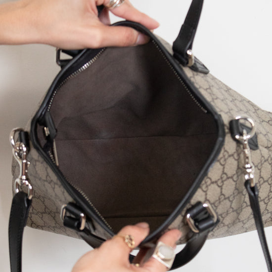 A close-up of a beige and black monogram handbag being held open by two hands against a plain white background.