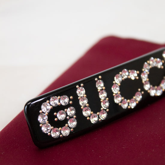 Black rectangular hair clip with 'GUCCI' in rhinestones on a red surface