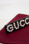 Black rectangular hair clip with 'GUCCI' in rhinestones on a red surface