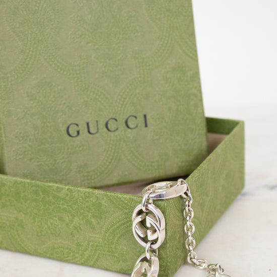 A silver-tone Gucci chain bracelet with interlocking G pattern links, displayed in front of its green Gucci gift box on a light marble surface.