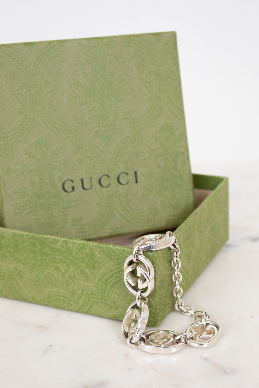 A silver-tone Gucci chain bracelet with interlocking G pattern links, displayed in front of its green Gucci gift box on a light marble surface.