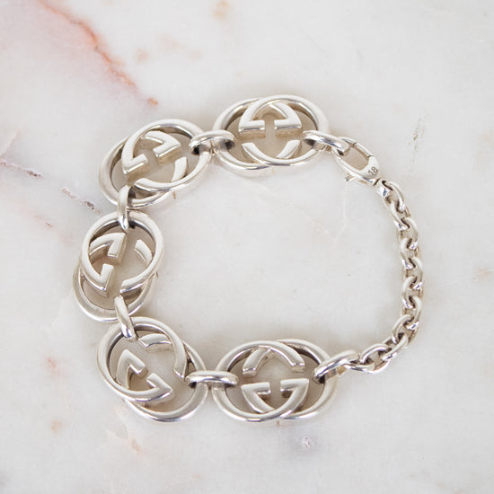 A silver-tone Gucci bracelet featuring interlocking GG logo links and a chain link closure, displayed on a white marble surface.