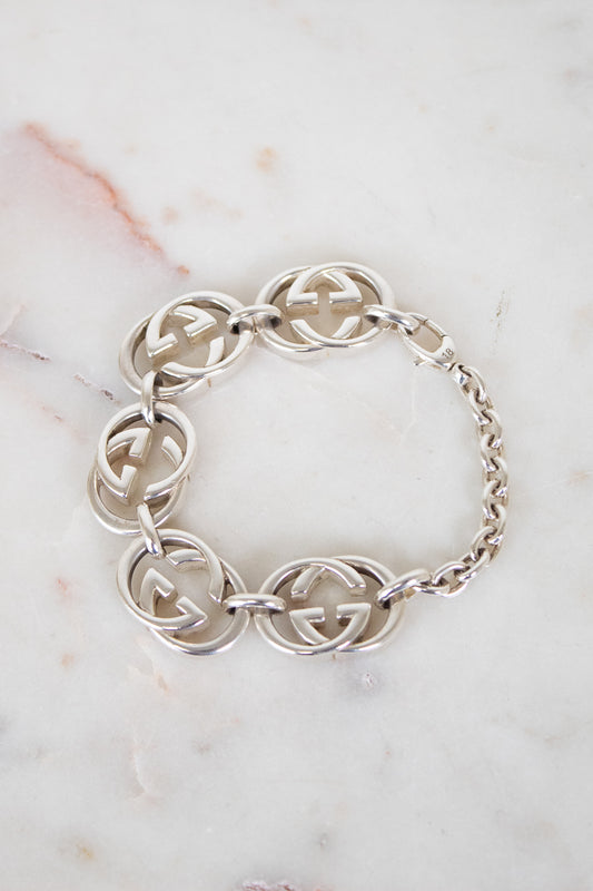 A silver-tone Gucci bracelet featuring interlocking GG logo links and a chain link closure, displayed on a white marble surface.