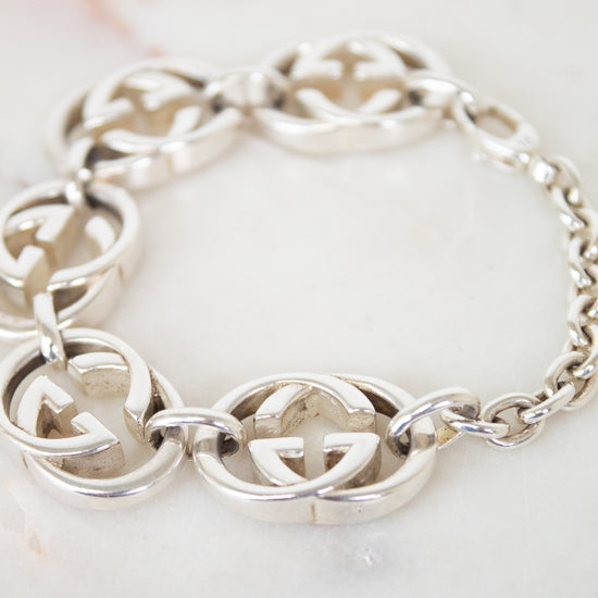 A silver-tone interlocking GG Gucci chain bracelet with oval chain links, displayed on a light marble surface. The brand is identified as Gucci due to the distinctive interlocking G logo pattern.