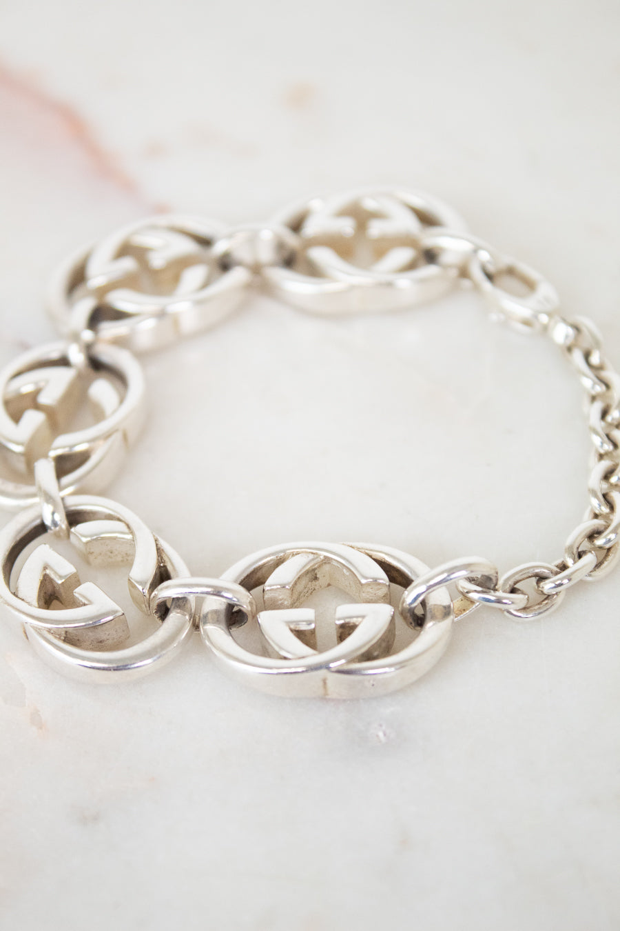 A silver-tone interlocking GG Gucci chain bracelet with oval chain links, displayed on a light marble surface. The brand is identified as Gucci due to the distinctive interlocking G logo pattern.