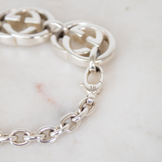 Close-up of a silver chain bracelet with interlocking double-G logo links and a lobster clasp, resting on a light marble surface. The Gucci brand is identified by the distinctive interlocking G logo and the '18' hallmark stamped on the clasp.