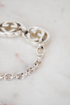 Close-up of a silver chain bracelet with interlocking double-G logo links and a lobster clasp, resting on a light marble surface. The Gucci brand is identified by the distinctive interlocking G logo and the '18' hallmark stamped on the clasp.