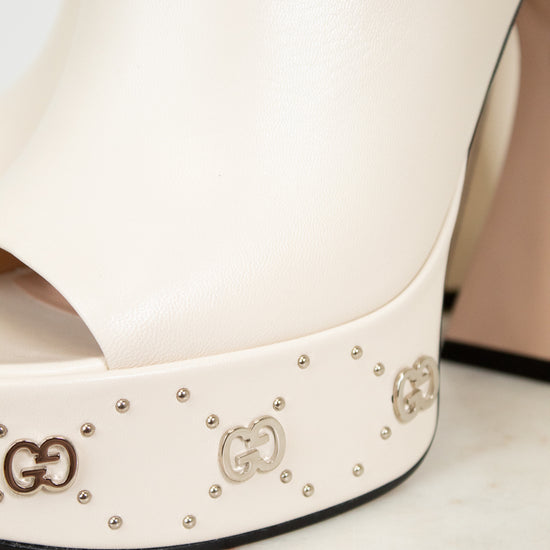 White platform high-heeled shoe with decorative logos and embellishments on a marble surface.