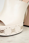 White platform high-heeled shoe with decorative logos and embellishments on a marble surface.