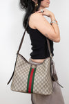 A woman wearing a black tank top and beige pants carries a beige Gucci monogram shoulder bag with brown leather trim and a green and red web stripe on a plain light gray background.