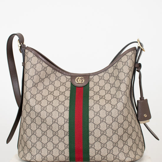 A Gucci monogram shoulder bag in beige GG canvas with the signature green and red web stripe down the center, dark brown leather trim and strap, and a gold-tone GG logo clasp, displayed on a white marble surface against a white background.