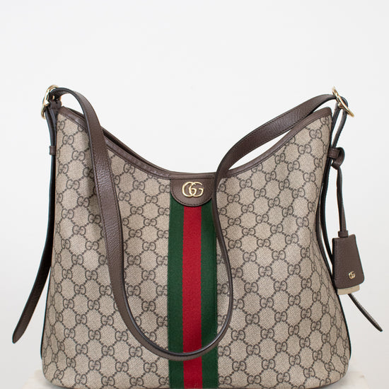Gucci GG Supreme shoulder bag in beige with brown leather trim, a green and red web stripe down the center, and a gold-tone GG logo clasp, displayed on a white marble surface against a white background.