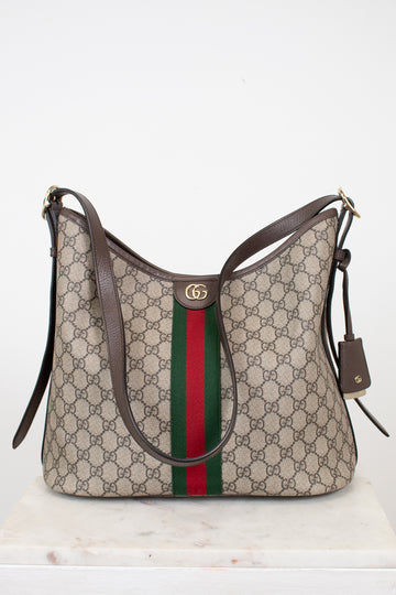 Gucci GG Supreme shoulder bag in beige with brown leather trim, a green and red web stripe down the center, and a gold-tone GG logo clasp, displayed on a white marble surface against a white background.