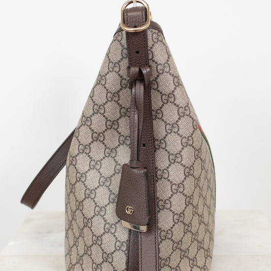 A Gucci shoulder bag in beige and brown GG Supreme canvas with dark brown leather trim and a gold-tone buckle, shown from the side on a light marble surface against a white background.