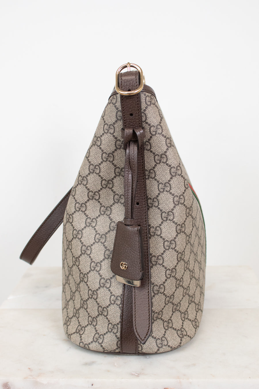 A Gucci shoulder bag in beige and brown GG Supreme canvas with dark brown leather trim and a gold-tone buckle, shown from the side on a light marble surface against a white background.