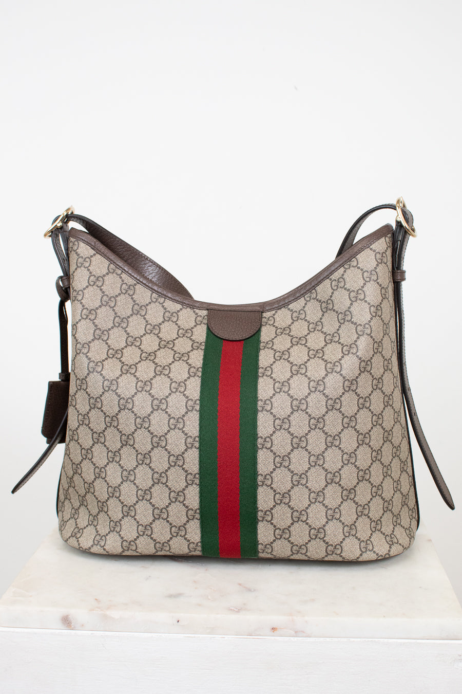 Gucci GG Supreme canvas hobo bag with brown leather trim and the signature green and red web stripe, displayed on a white marble surface against a white background.