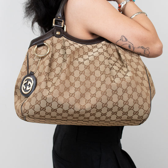 Person standing in front of white background in a black outfit holding beige handbag with brown Gucci monogram print. 
