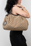 Person standing in front of white background in a black outfit holding beige handbag with brown Gucci monogram print. 