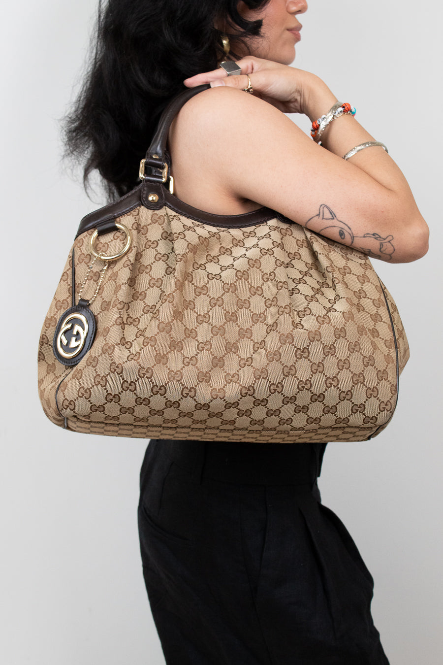 Person standing in front of white background in a black outfit holding beige handbag with brown Gucci monogram print. 
