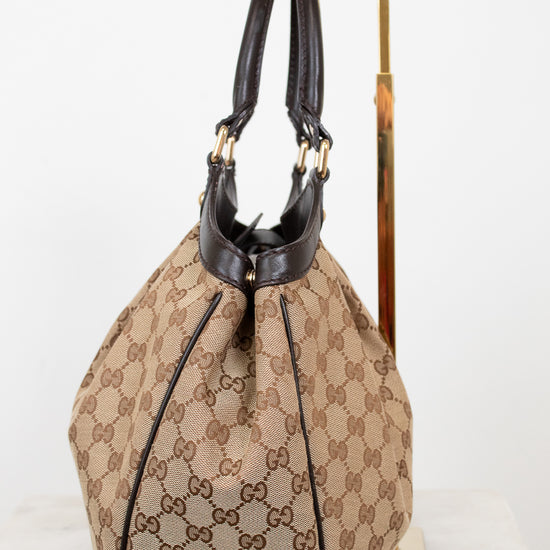 Side view of beige handbag with brown Gucci monogram with brown handles on a white background