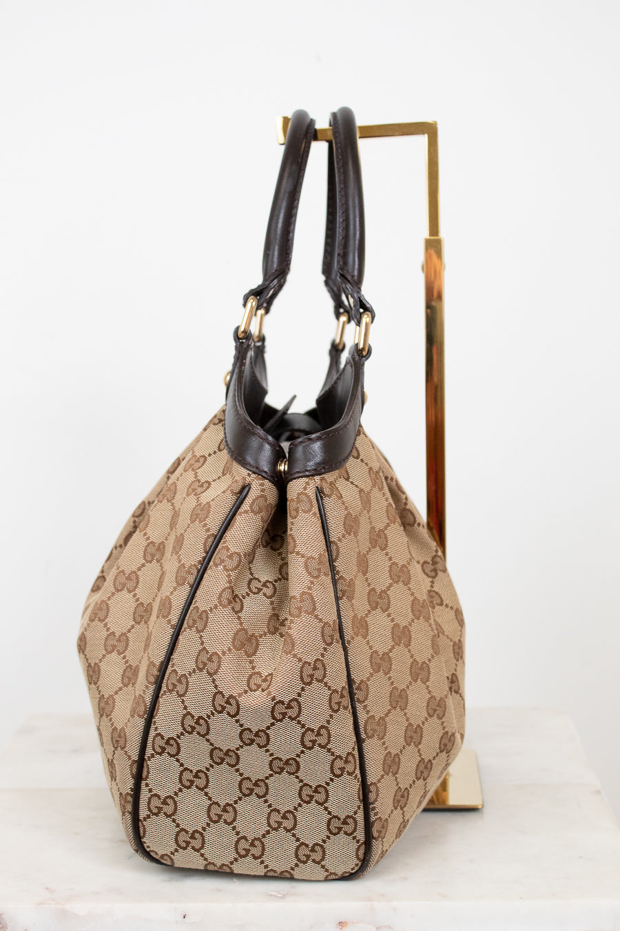 Side view of beige handbag with brown Gucci monogram with brown handles on a white background