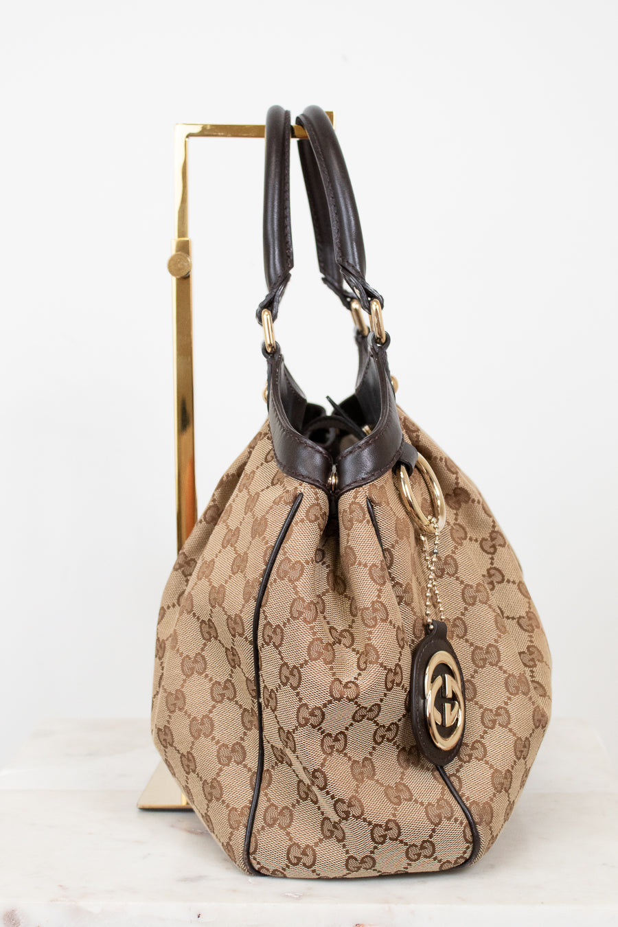 Side view of beige handbag with brown Gucci monogram with brown handles on a white background