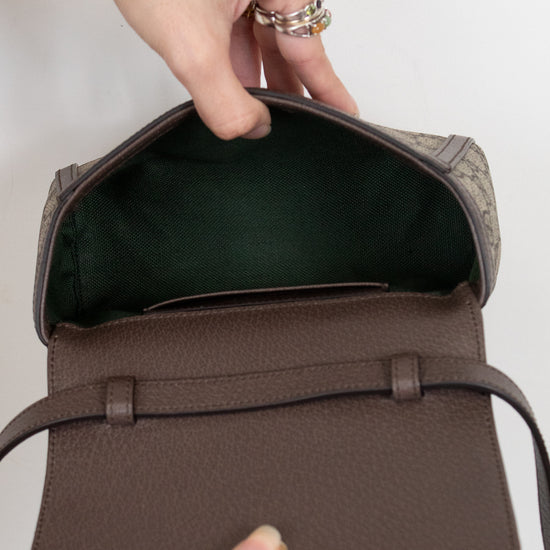 A close-up of a brown textured leather handbag being opened by hands wearing gold rings, showing a dark interior lining and a patterned exterior fabric.