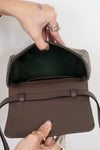 A close-up of a brown textured leather handbag being opened by hands wearing gold rings, showing a dark interior lining and a patterned exterior fabric.