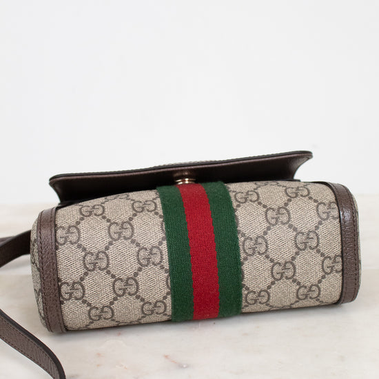 Bottom view of a Gucci crossbody bag made of beige GG Supreme coated canvas with brown leather trim and a green and red web stripe, resting on a white marble surface against a white background.