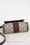 Bottom view of a Gucci crossbody bag made of beige GG Supreme coated canvas with brown leather trim and a green and red web stripe, resting on a white marble surface against a white background.