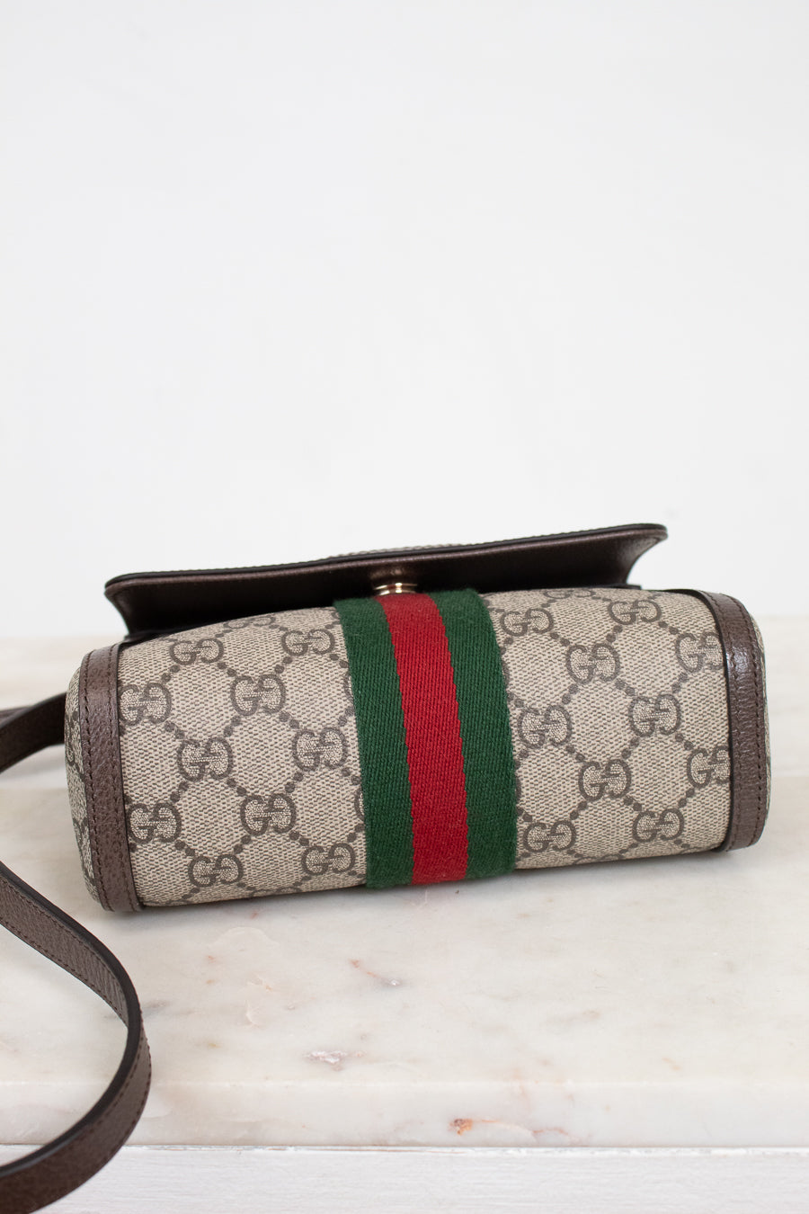 Bottom view of a Gucci crossbody bag made of beige GG Supreme coated canvas with brown leather trim and a green and red web stripe, resting on a white marble surface against a white background.