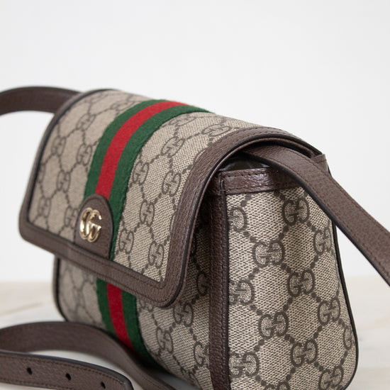 Gucci GG Supreme coated canvas shoulder bag with brown leather trim, a green and red web stripe, and gold-tone hardware, shown on a light marble surface against a white background.