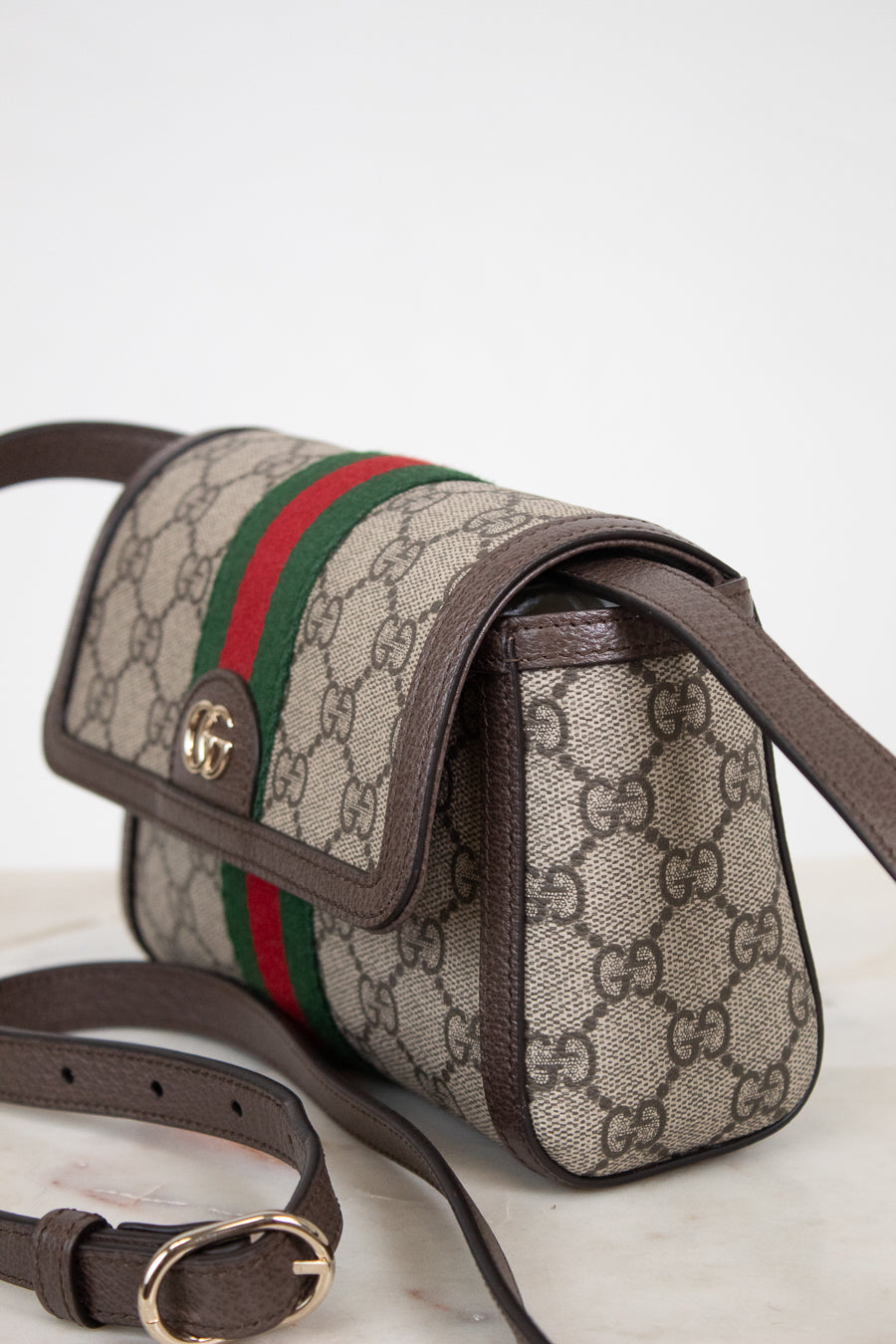Gucci GG Supreme coated canvas shoulder bag with brown leather trim, a green and red web stripe, and gold-tone hardware, shown on a light marble surface against a white background.