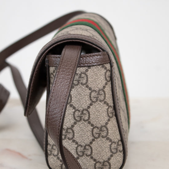 A close-up of the side view of a Gucci shoulder bag featuring the classic GG Supreme coated canvas in beige and brown, with dark brown leather trim and a signature green and red web stripe.