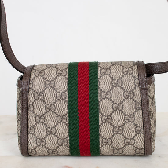 A small Gucci crossbody bag in beige GG Supreme coated canvas with brown leather trim and a signature green and red web stripe down the center, shown on a light marble surface against a white background.