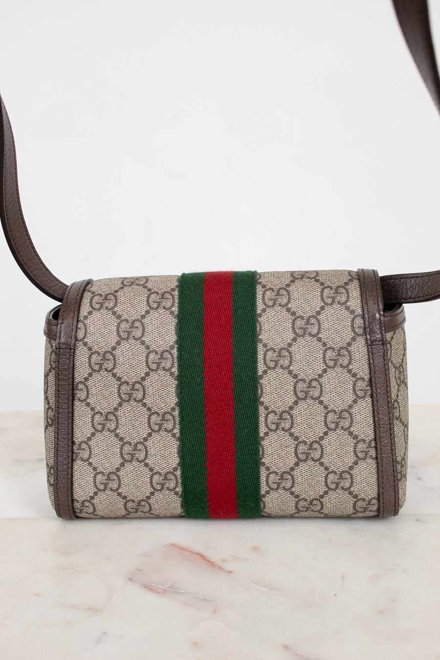 A small Gucci crossbody bag in beige GG Supreme coated canvas with brown leather trim and a signature green and red web stripe down the center, shown on a light marble surface against a white background.