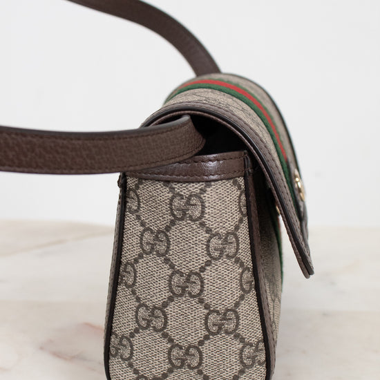 A close-up of a Gucci crossbody bag featuring the classic GG Supreme coated canvas in beige and brown, dark brown leather trim, and the signature green and red web stripe detail on the strap.