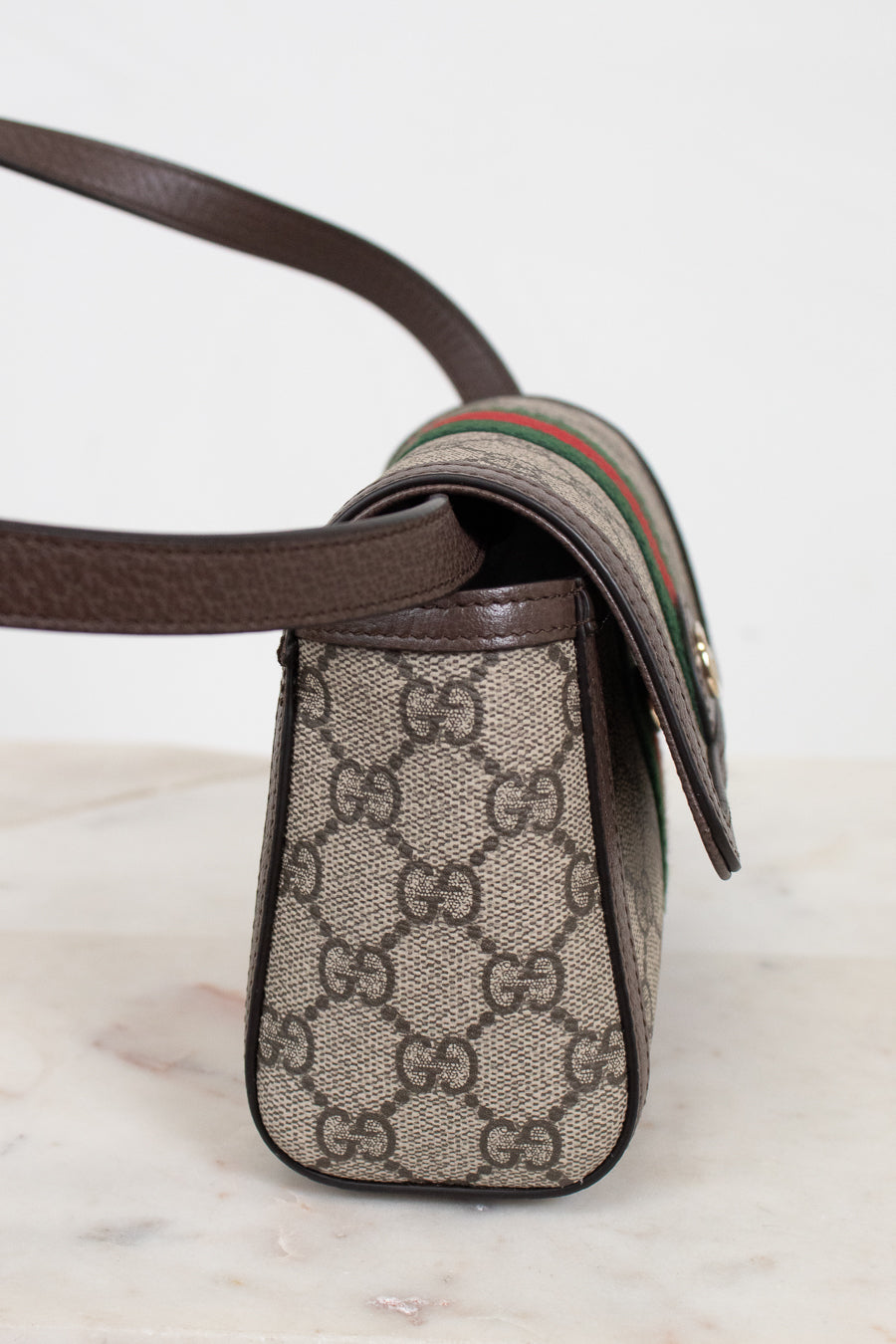 A close-up of a Gucci crossbody bag featuring the classic GG Supreme coated canvas in beige and brown, dark brown leather trim, and the signature green and red web stripe detail on the strap.