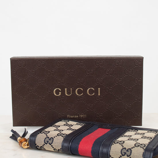 Brown box with branded 'GUCCI' with navy and monogrammed zip around wallet with red and navy striped center detail with gold and bamboo zipper laying against a marble background