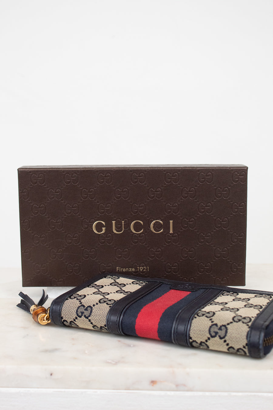Brown box with branded 'GUCCI' with navy and monogrammed zip around wallet with red and navy striped center detail with gold and bamboo zipper laying against a marble background
