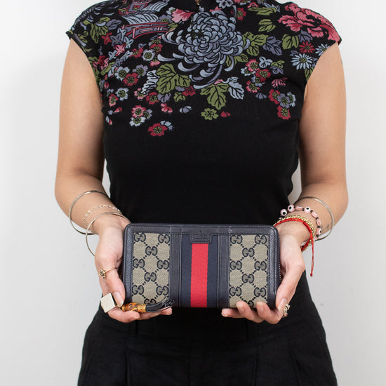 Woman wearing embroidered black top holding 'Gucci' monogrammed wallet with red and navy center stripe detail and standing against a plain background