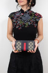 Woman wearing embroidered black top holding 'Gucci' monogrammed wallet with red and navy center stripe detail and standing against a plain background