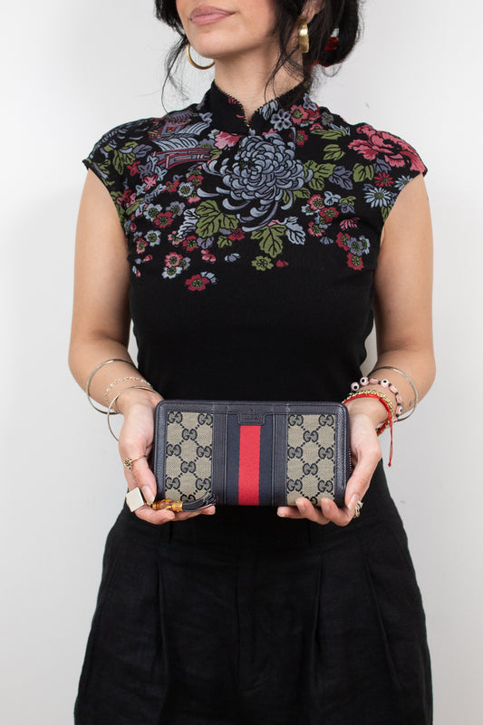 Woman wearing embroidered black top holding 'Gucci' monogrammed wallet with red and navy center stripe detail and standing against a plain background