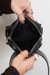 Person opening a black leather handbag with zippers on a neutral background