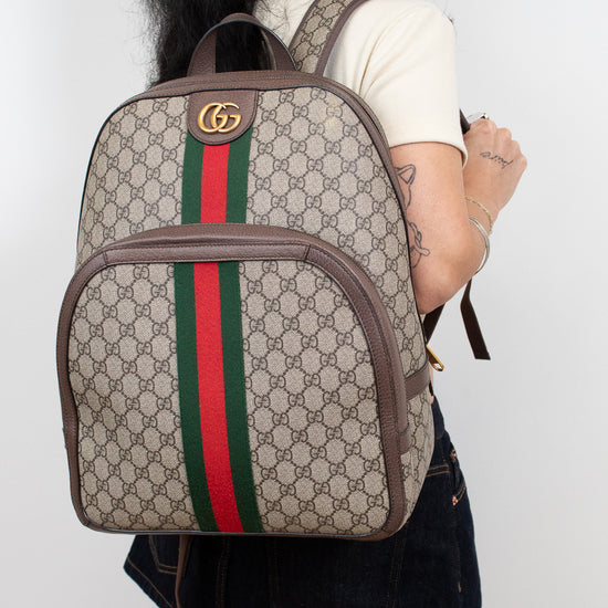 A person shown from the back wearing a beige Gucci backpack with the classic GG monogram pattern, brown leather trim, and the signature green and red web stripe down the center.